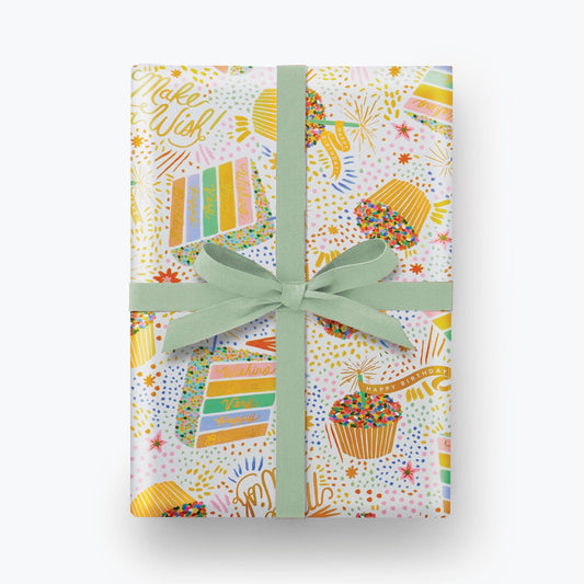 Birthday Cake Wrapping Paper shown with a blue bow. Illustration on wrapping paper features cupcakes and slices of colorful layer cake with lots of sprinkles. Multiple areas of text read "Make a WIsh!" and "Wishing you a very Happy Birthday" in gold script.