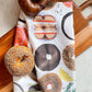 Tea towel with different types of bagels on it. It is laid out on a cutting board next to a sesame bagel and a pumpernickel bagel.  