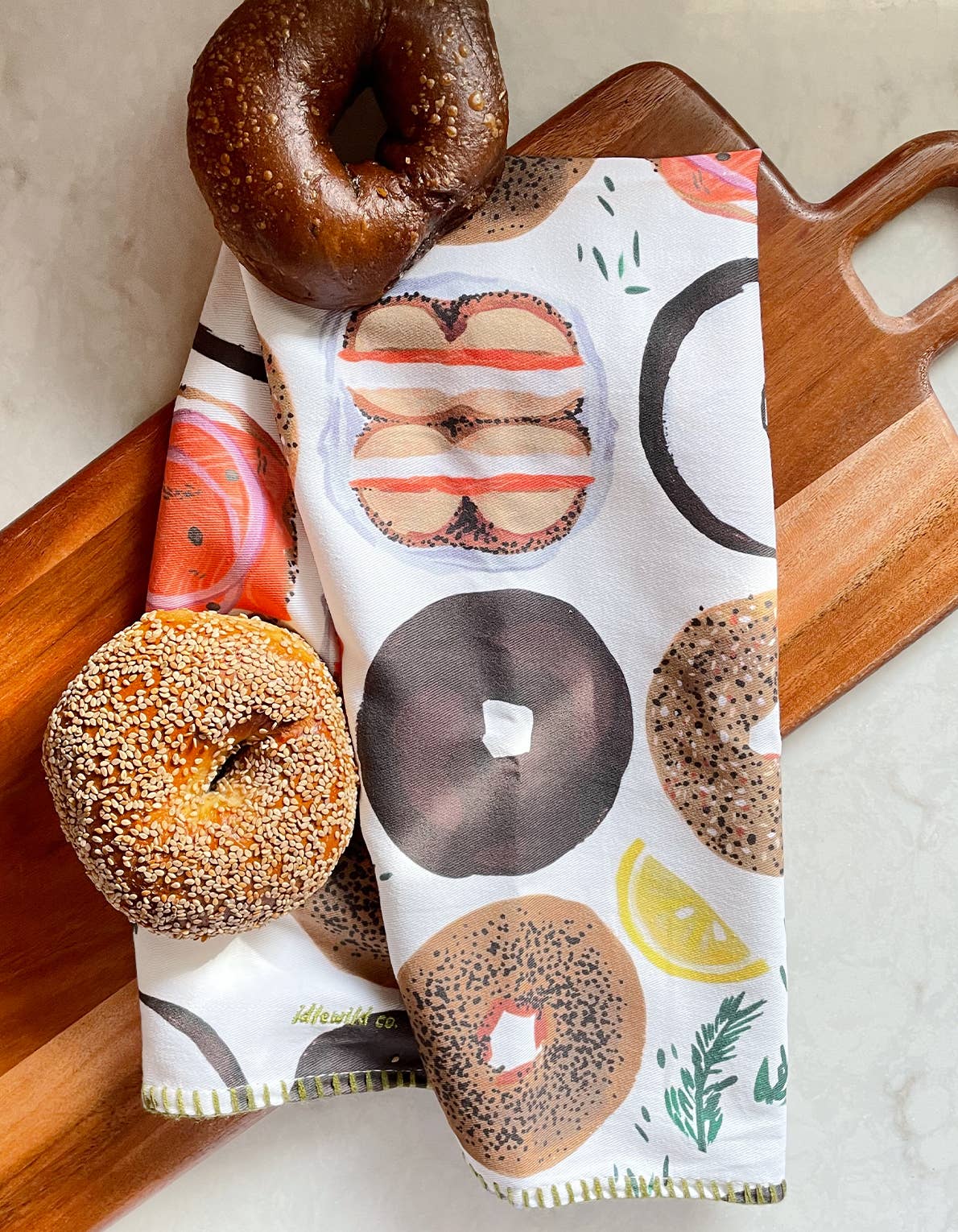 Tea towel with different types of bagels on it. It is laid out on a cutting board next to a sesame bagel and a pumpernickel bagel.  