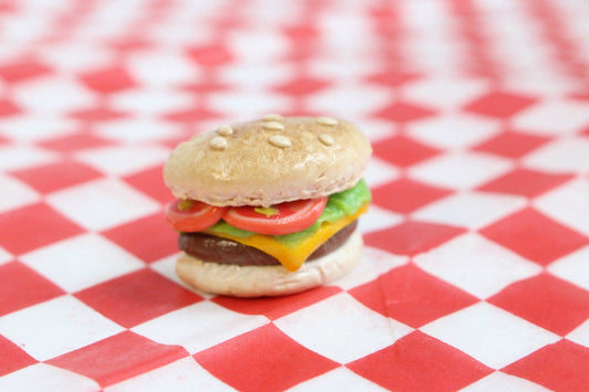 Single tiny cheeseburger magnet pictured on a mini red and white checkered tablecloth background. Cheeseburger is hand-sculpted and depicts a pale bun with tiny seeds, brown burger, melted American cheese, lettuce and tomato.