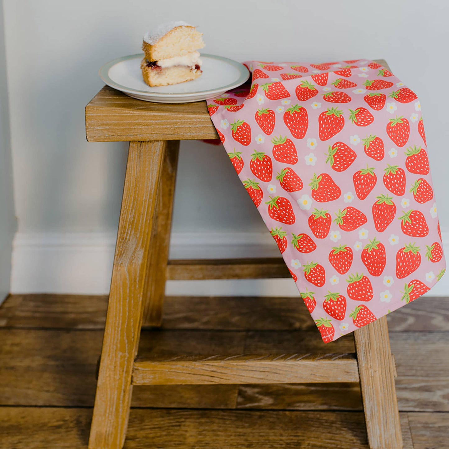 Photo of strawberry tea towel draped over short wooden stool. Also on top of the stool is a slice of cake on a white ceramic plate.