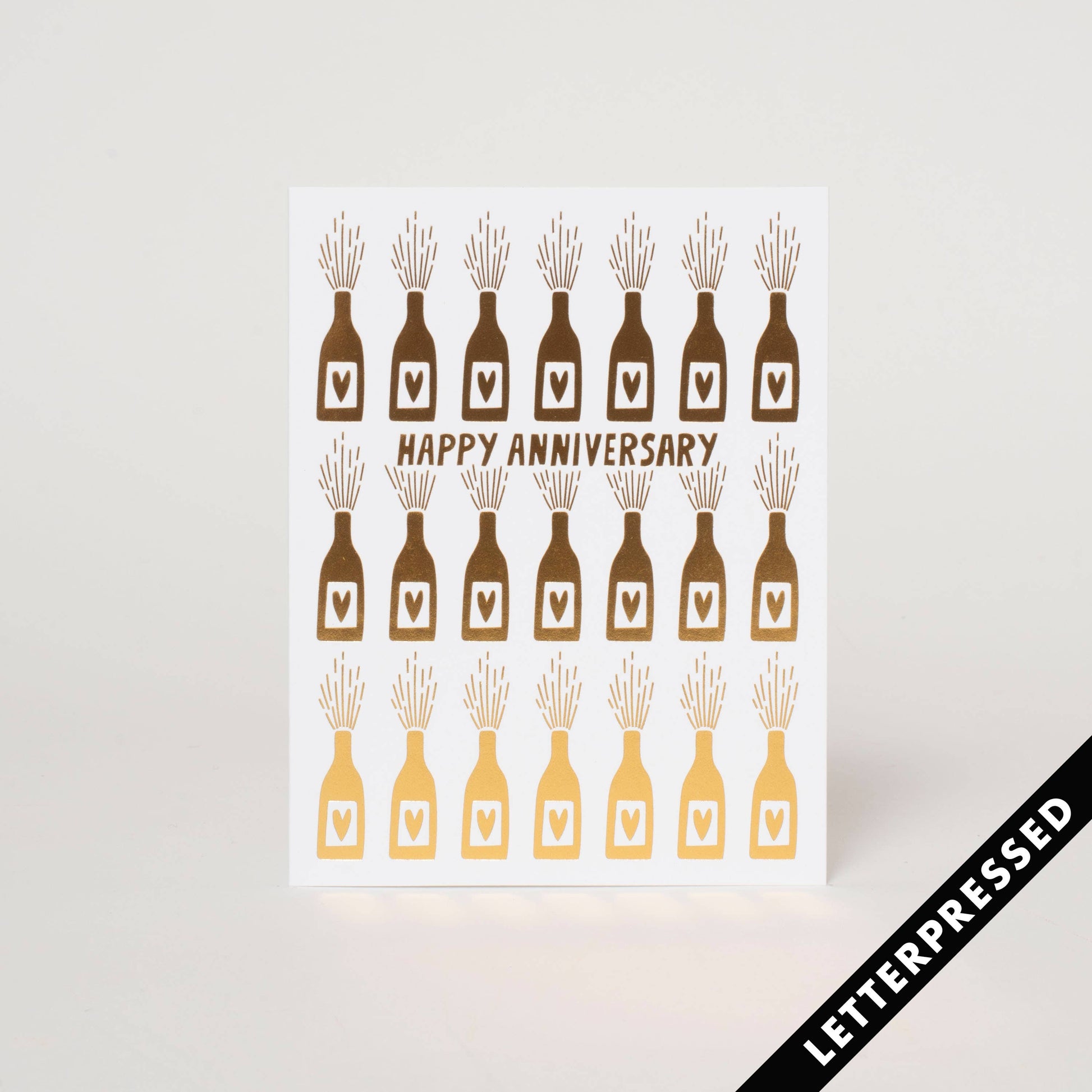 Greeting card printed in metallic ink. Text "Happy Anniversary" surrounded by wine bottles with hearts on each label, popping open in celebration.