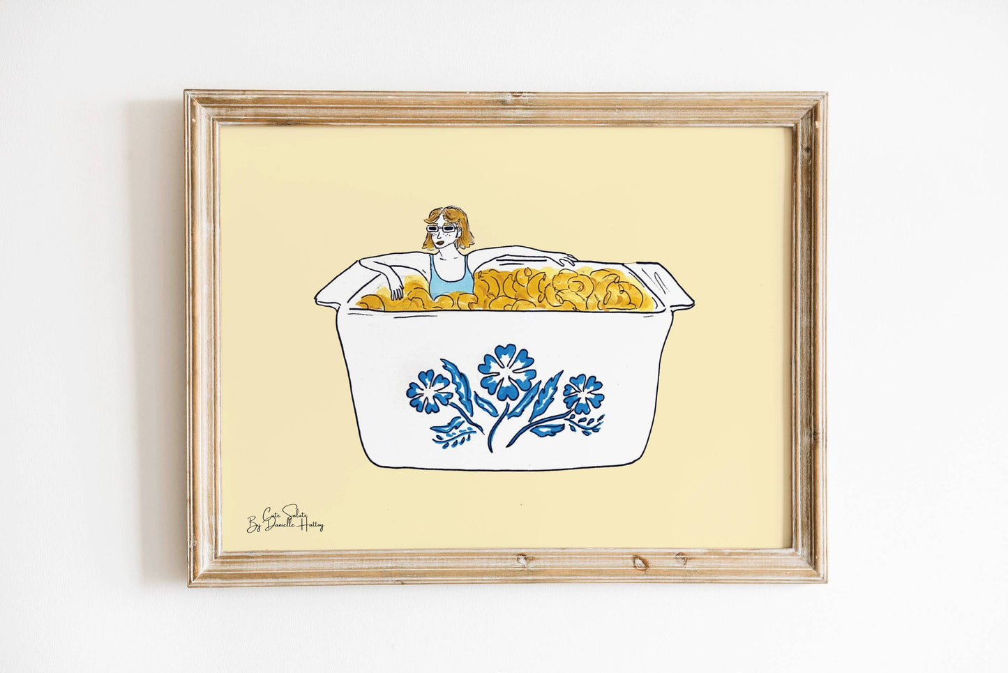 Wall print featuring an illustration of a girl sitting in a vintage CorningWare dish of Mac and Cheese with a pale yellow background. Shown in light colored wood frame.