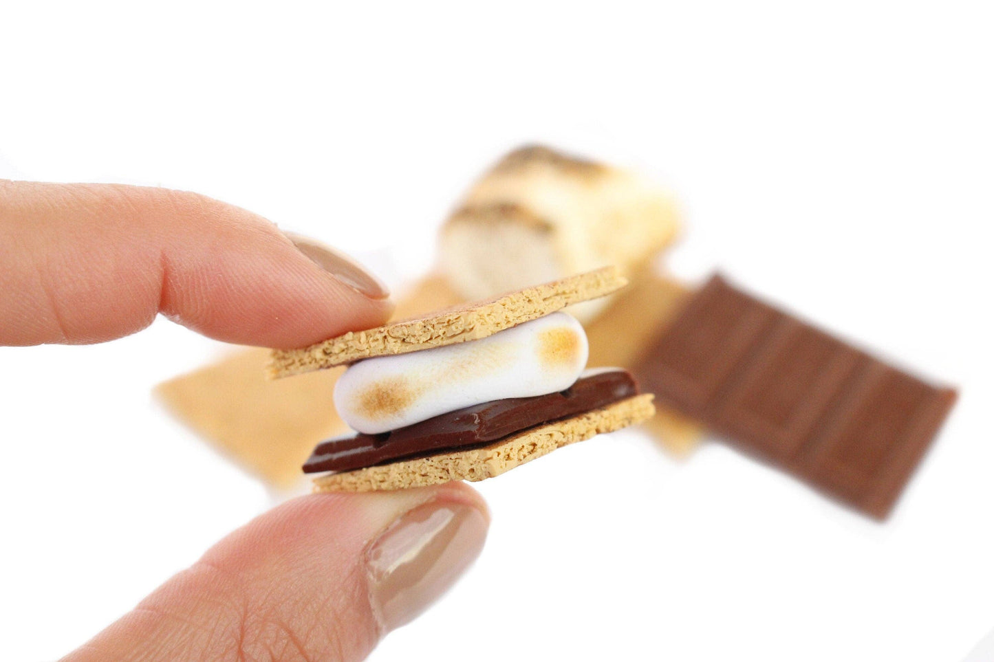 Magnet shaped like a single s'more - toasted marshmallow on chocolate, sandwiched between two triangles of graham cracker. Shown held between two fingers for size comparison.
