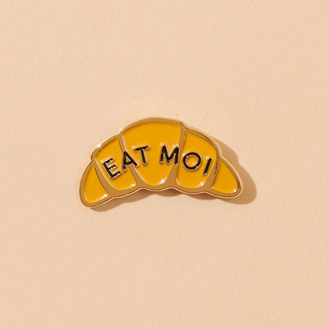 Enamel pin in the shape of a croissant that says "EAT MOI"
