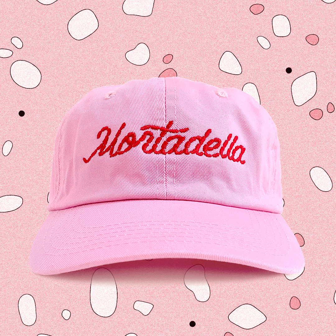 Mortadella dad hat -- collaboration between Mister Parmesan and Marianna Fierro; Pink baseball cap with Mortadella stitched on the front in red. Background is mostly pink . 